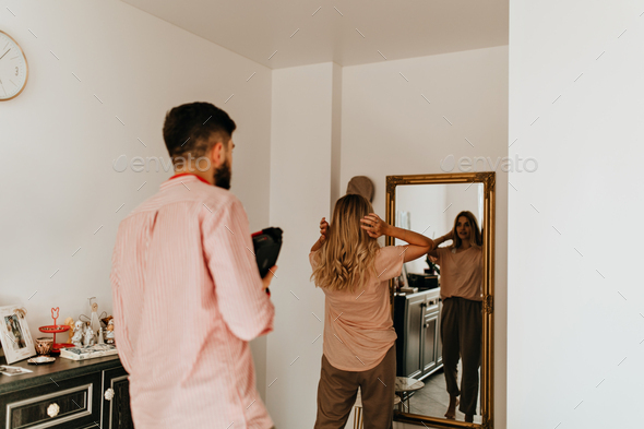 Man in pink shirt with camera in his hands looks at how his beloved wife preens in front of mirror