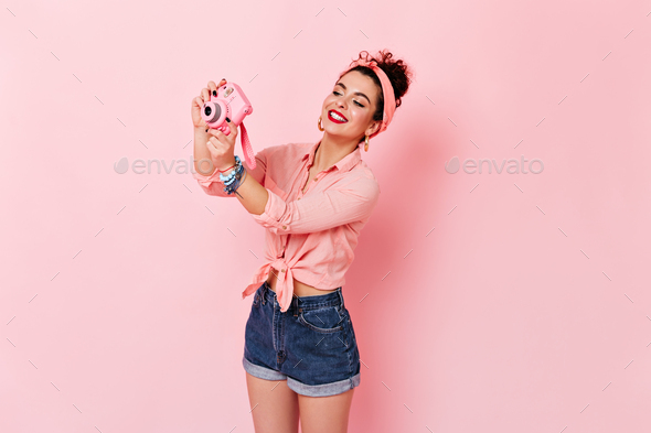 Curly woman in headband, pink blouse and denim shorts makes photo on mini camera on pink background
