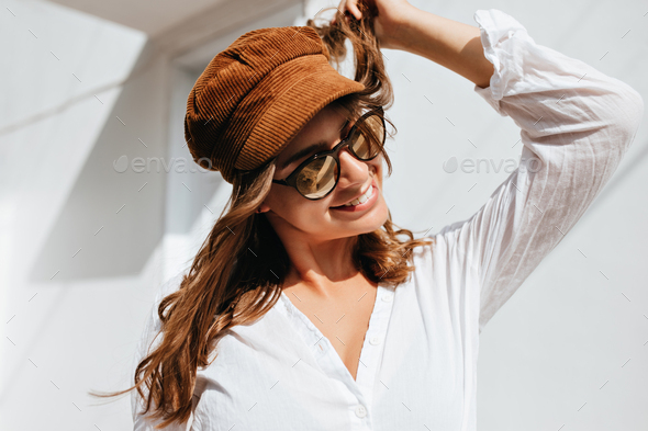 Young woman plays with her hair and smiles. Girl dressed in white blouse, brown hat and sunglasses