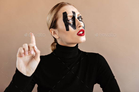 Blue-eyed girl with face art in form of word no shows index finger, posing in black sweater on beig