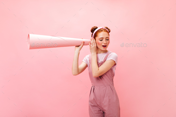 Young mischievous red-haired girl in pink jumpsuit eavesdrops using pink tube