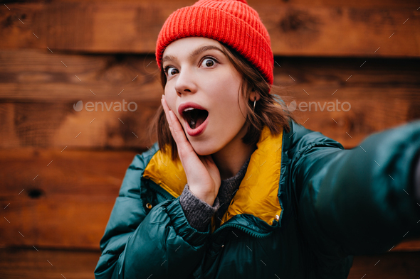 Funny woman in stylish winter clothes stares in surprise at camera on wooden background. Short-hair