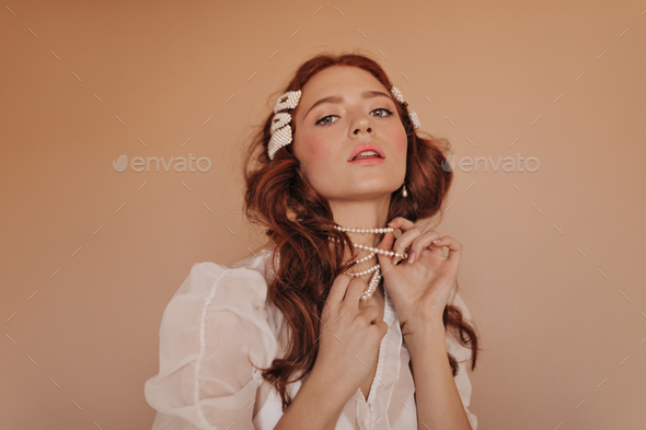 Pink-cheeked redhead girl plays with her pearl necklace. Lady in classic white blouse looking down