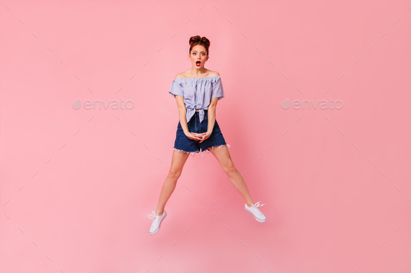 Full length view of shocked woman in denim shorts and blouse. Studio shot of amazed pinup girl jump