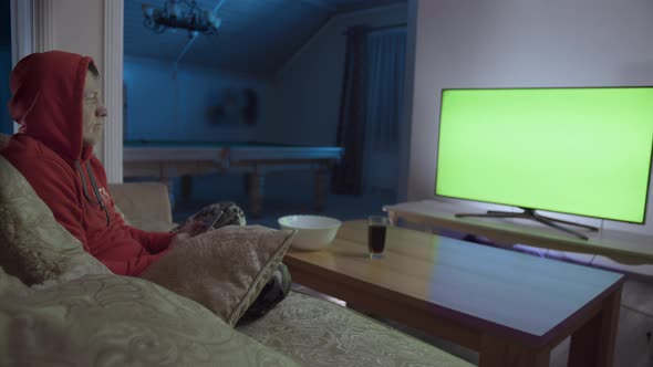 A Man Sits in the Evening on a Couch with a Mobile Phone in Front of a Green Screen TV alt