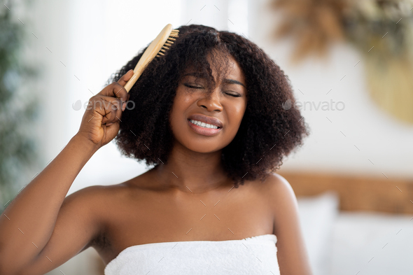 Frustrated millennial black woman trying to brush her tangled hair at home. Domestic hairdressing