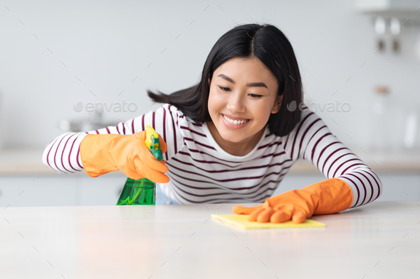 Smiling asian lady cleaning kitchen table with spray and cloth