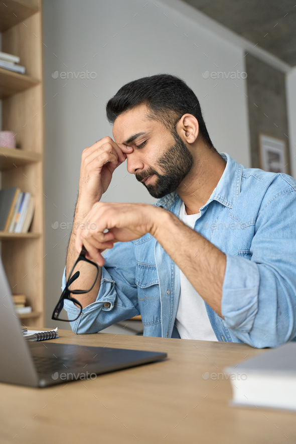 Stressed indian student rubbing nose bridge sitting at desk. Vertical shot.