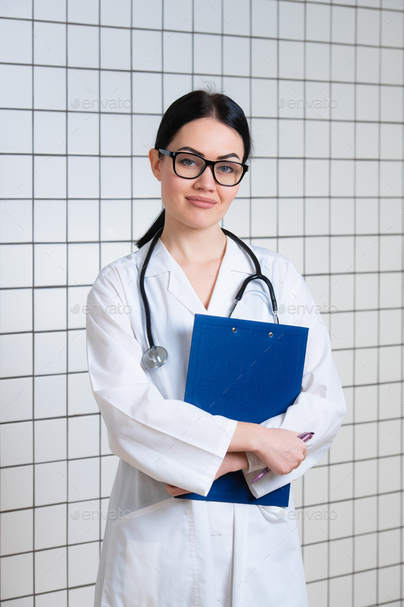 Young beautiful female doctor in white surgical coat with black stethoscope and blue paper holder in