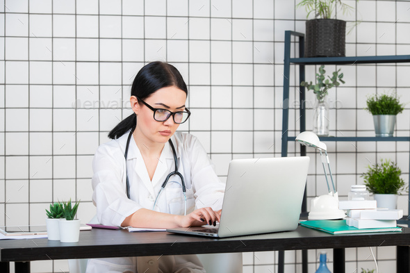 Young smiling woman sitting at desk, working on computer with medical documents in light office in - Stock Photo - Images