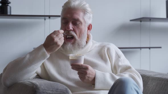 A Handsome Elderly Man with a Gray Beard Eats Yogurt Holding a Spoon Close To His Mouth alt