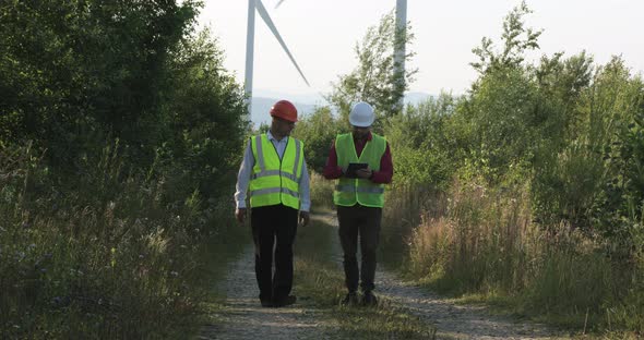 Two Young Engineers Walk on a Dirt Road Looking at a Tablet Showing Statistics of Air Energy and alt