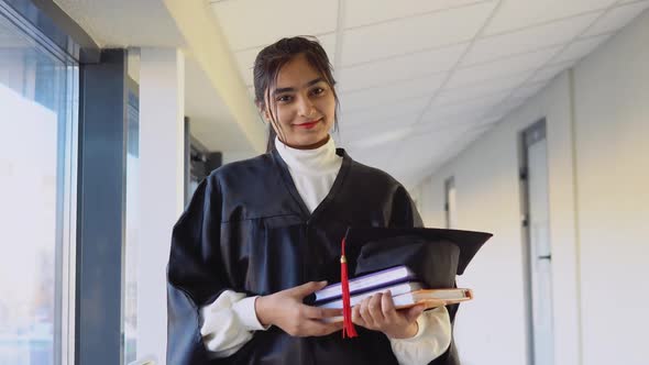 Pakistani Female Graduate in Mantle Stands with a Books in Her Hands and Smiles alt