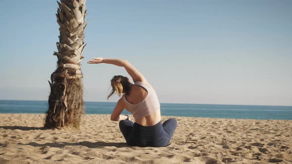 Woman in Sportswear Sits on the Beach at Sunny Day Enjoying Life and Doing Yoga and Stretching alt
