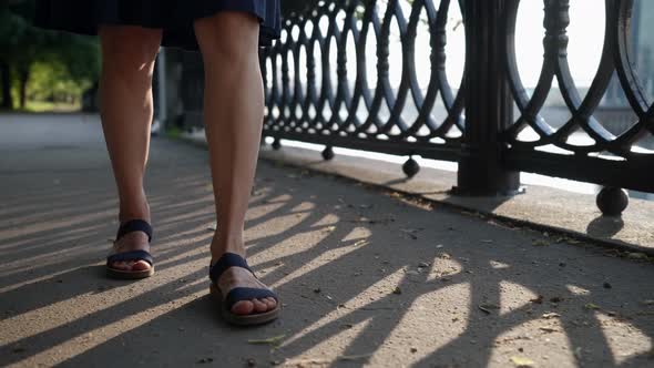 Female Feet in Black Sandals Walk on the Sandy Road Along the Openwork Lattice alt