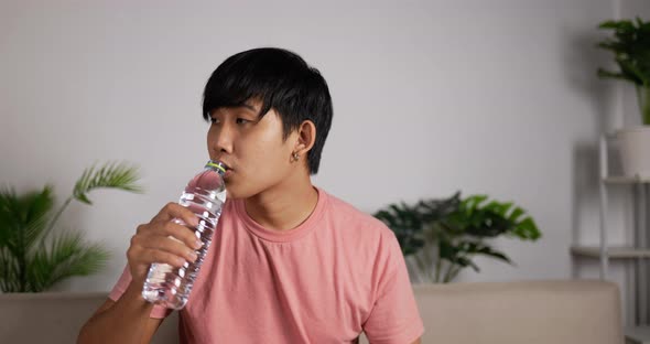 Tired Asian man sitting on a couch drink chilly fresh water from a bottle. alt
