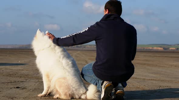 A man with a dog sits on the shore of the lake. A man is stroking a white fluffy dog Samoyed alt