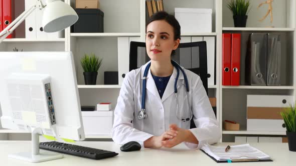 Doctor in White Lab Coat Sitting at Workplace in Bright Office and Looking at Camera