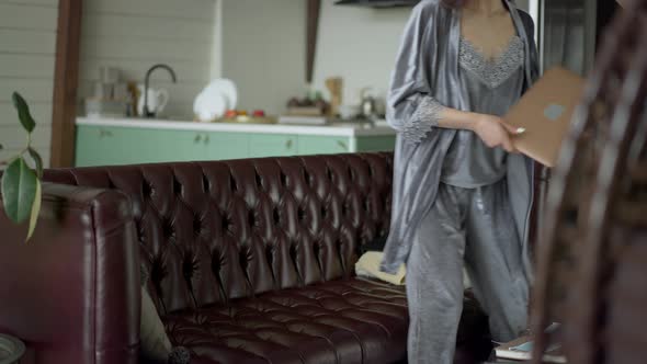 Young Charming Asian Woman in Sleepwear Entering Living Room Sitting Down on Couch and Opening alt