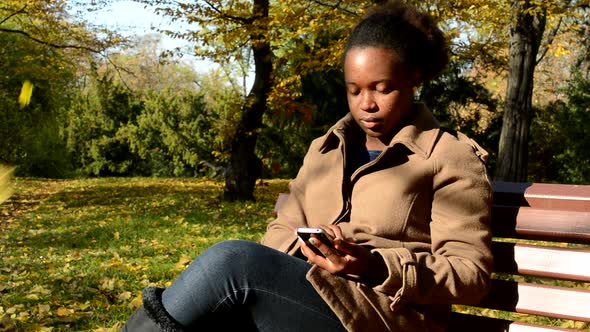Young Beautiful African Serious Girl Sits on Bench in Woods and Works on Phone alt