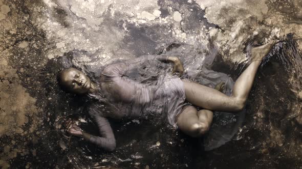 A Woman in a Dress with Dyed Golden Skin and Hair Lies in the Water Slowly Moving and Posing alt