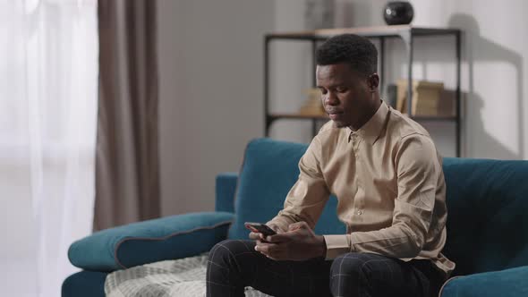 Young Afroamerican Man is Using Smartphone Sitting on Couch in Apartment Male Internet User Viewing alt