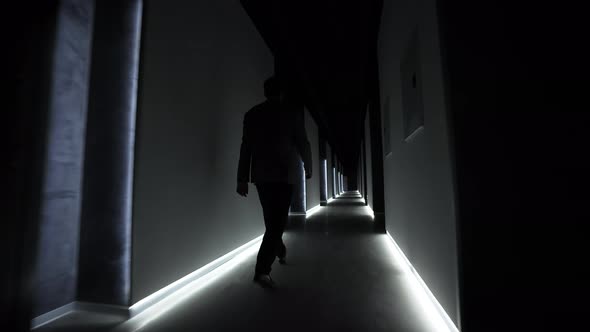 Man in a Jacket Walks Along a Long Dark Corridor with Lights on the Floor