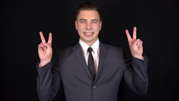 Young Businessman Shows a Victory Sign with Two Hands. Man in a Black Suit on a Black Background alt