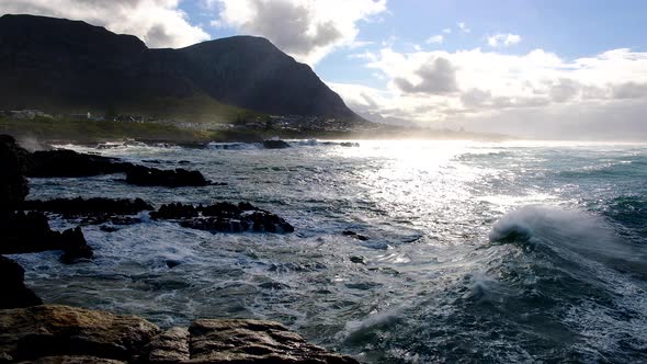 Wave reflecting beautiful morning light as it rolls past onto rocky coastline, with mountains in bac alt