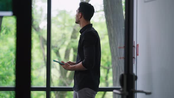 Bearded Thoughtful Middle Eastern Young Man Scrolling Tablet Looking Out the Window in Office alt