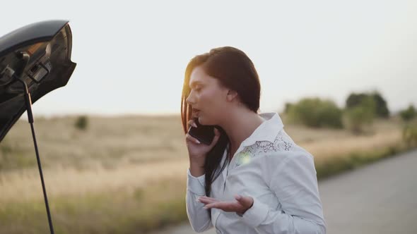 Perplexed Business Lady Looking Under Car Hood and Talking on Phone on Sunset alt