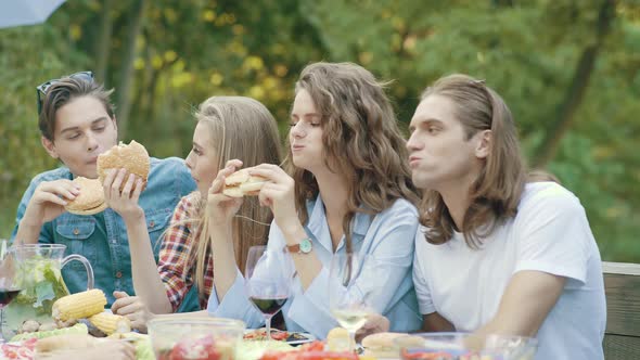 Friends Eating Burgers Sitting At Dinner Table At Outdoor Party alt