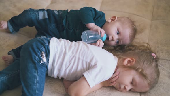 Funny Baby Looks at Sleeping Sister Drinking Water on Bed alt