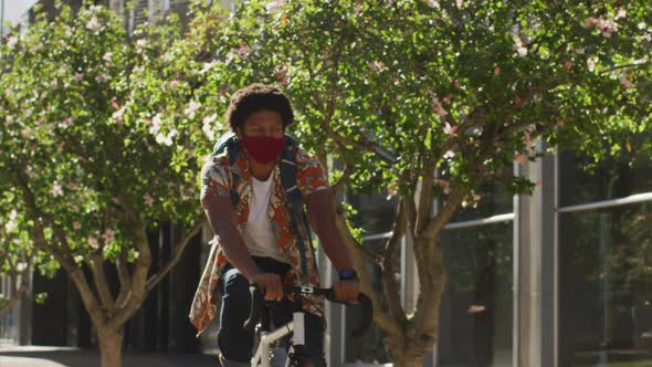 African american man in city, wearing face mask riding bicycle in street alt