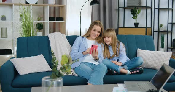 Sisters Sitting Together on Soft Couch in Contemporary Apartment and Using Smartphone alt