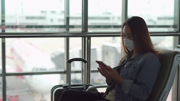 Young asian woman wearing sergical mask using smart phone while waiting for her flight