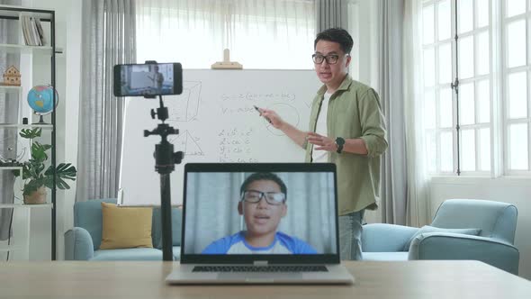 Teacher With Glasses Shoots Video By Smartphone While Having Video Call On Laptop And Teaching Math alt