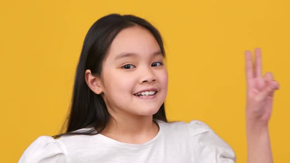 Close Up Portrait of Adorable Little Asian Girl Showing V Sign to Camera and Smiling Widely Orange alt