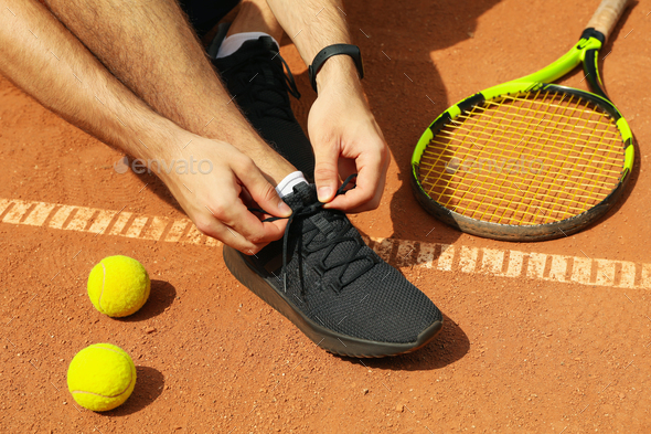 Man ties his shoelaces on clay court with racket and balls