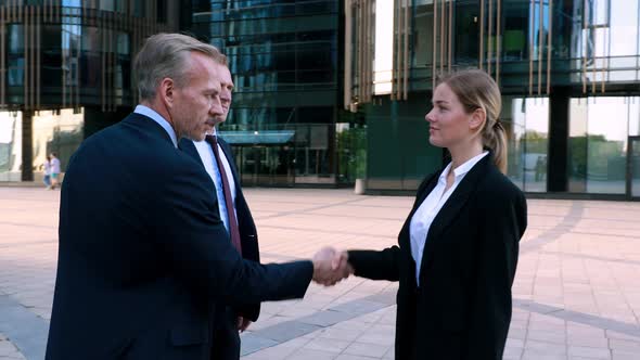 Woman Walking To Businessmen and Shaking Hands Looking at Camera Three Business Partners in Front