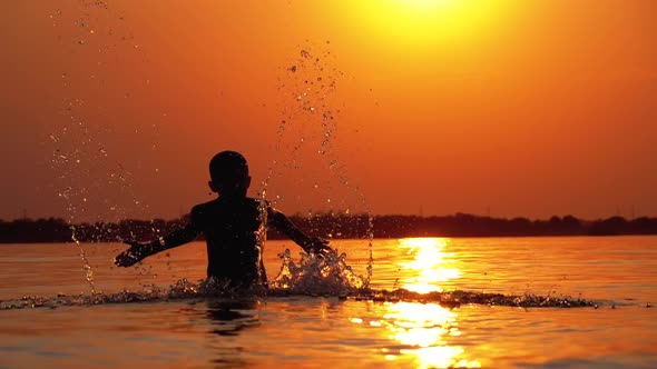 Silhouette of Boy at Sunset Raises Hands and Creating Splashes of Water. Slow Motion alt