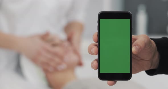 Man's Hand Holding Green Screen Smartphone on the Cosmetology Procedures Background alt