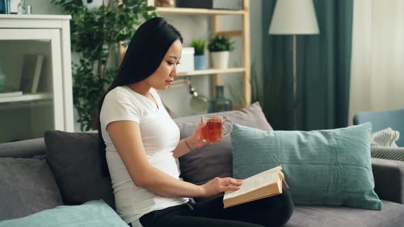 Good Looking Asian Woman Is Reading Book Enjoying Modern Good Looking Asian Woman Is Reading Book Enjoying Modern Literature Sitting On Sofa At Home Drinking Stock Footage