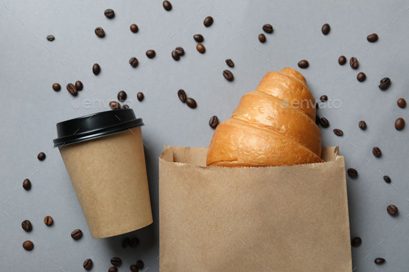 Paper bag with croissant, paper cup and coffee seeds on gray background