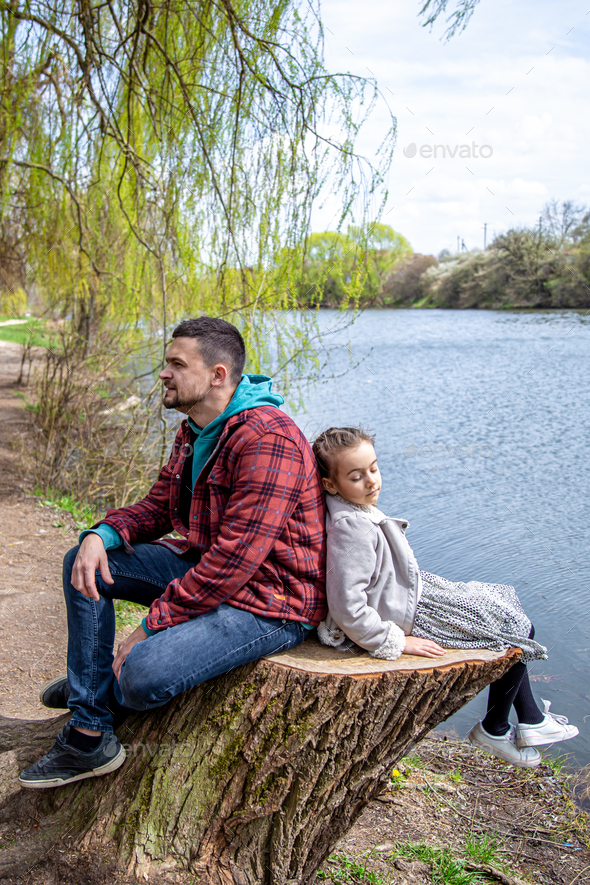 A little girl and dad are sitting on a tree stump by the river.