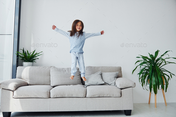 Jumping on the sofa. Cute little girl indoors at home alone. Child enjoying weekend
