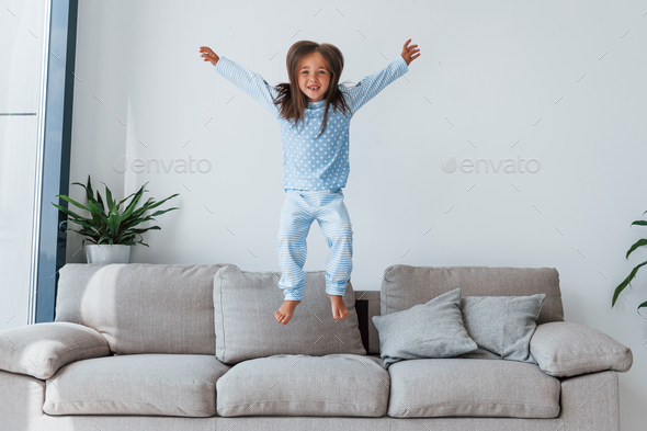 Jumping on the sofa. Cute little girl indoors at home alone. Child enjoying weekend
