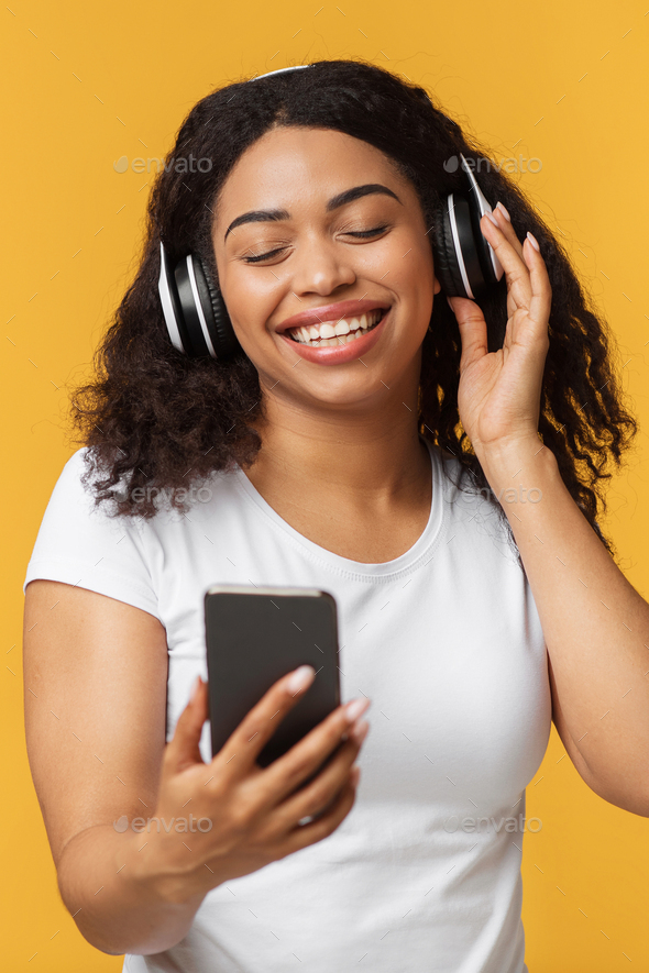 Happy black lady enjoying newest stereo headset and mobile music app, listening to music over yellow