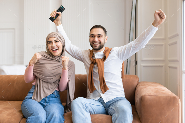 Emotional Muslim Couple Shaking Fists Watching TV At Home