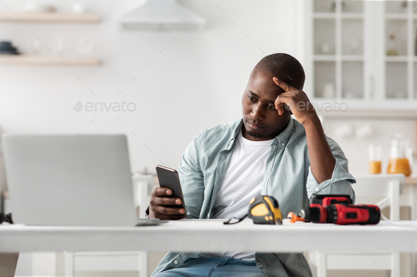 Upset african american man having problems with furniture instructions looking at phone, siting at